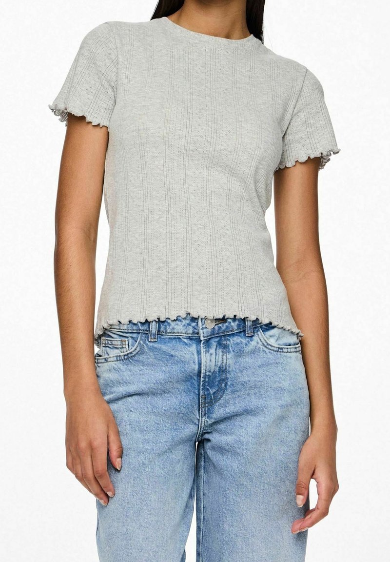 Femme portant un haut en maille côtelée gris clair à manches courtes avec des bords festonnés, associé à un jean en denim bleu clair taille haute.