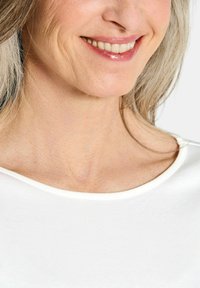 Witte stoffen top met een ronde halslijn, die een gladde textuur en subtiele stikseldetails laat zien. Huidtint zichtbaar rond de nek en kin.