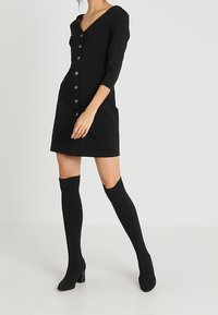 Mustapuku, jossa on V-pääntie, kolmannes hihat ja nappilista. Yhdistetty polven ylle ulottuviin mustiin sukkiin ja korkokenkiin. Liukas kangas.