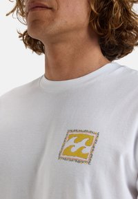 White cotton t-shirt featuring a small embroidered square patch with yellow wave patterns, bordered by a colorful decorative outline.