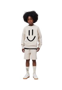 Beige színű pulóver fekete mosolygó arc mintával, bézs shorttal párosítva. Fehér sportcipő és térd fölé érő zokni, amelyen szintén mosolygó arcok vannak.