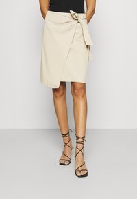 Jupe portefeuille beige avec un détail à nouer à la taille, avec une texture douce et un design à longueur genou ; assortie à des sandales noires à lanières.