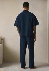 Sötétkék, rövidujjú, texturált ing és hozzá illő nadrág, amely laza szabást biztosít. Puha anyag semleges háttérrel.