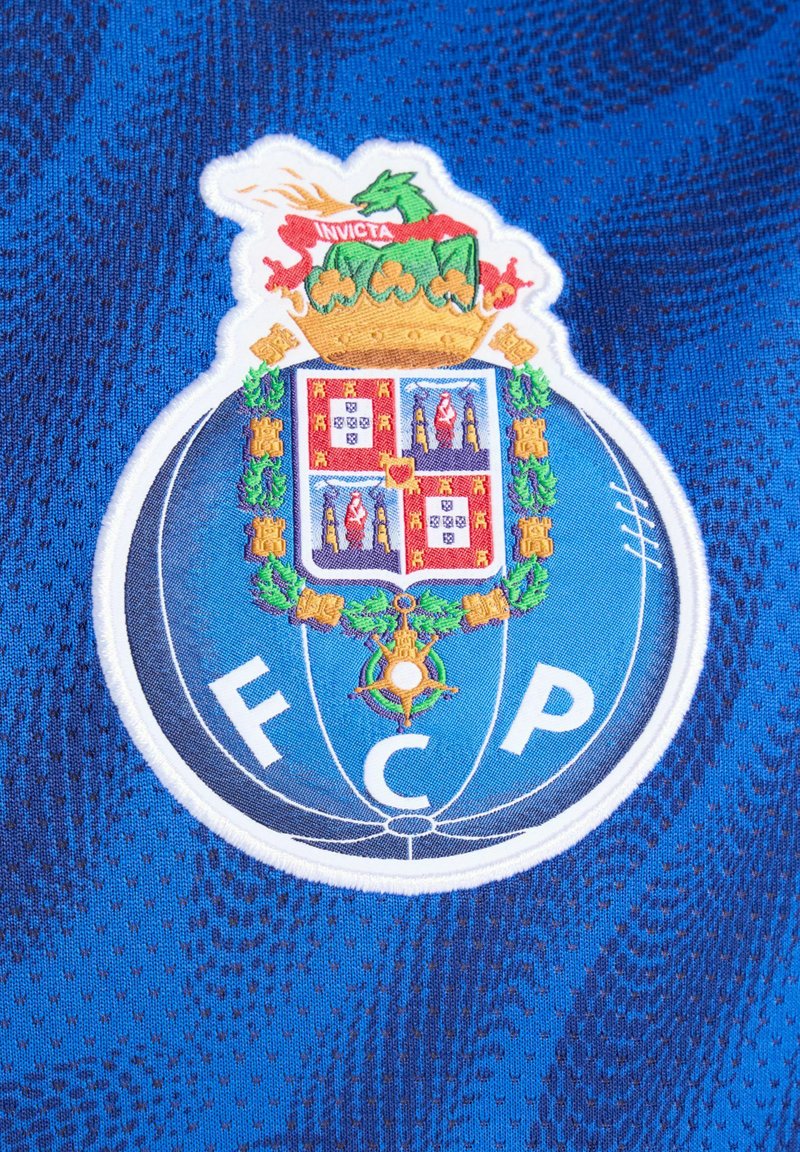 Blauw shirt met een gedetailleerd geborduurd logo. Het wapen bevat een kroon, een draak en kleurrijke kwartiervelden, met "FCP" aan de onderzijde.