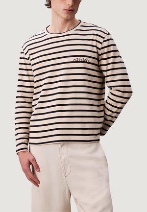 Joven vestido con una camisa de rayas horizontales beige y negra de manga larga con texto bordado y pantalones de color claro, con la mano en el bolsillo.