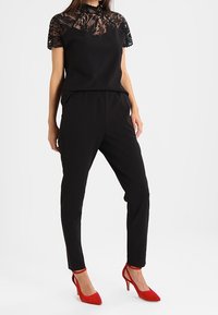 Top negro con superposición de encaje, combinado con pantalones negros ajustados y tacones rojos. Presenta un escote redondo y mangas cortas.