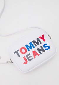 Borsa crossbody bianca con forma rettangolare, caratterizzata da un testo audace "TOMMY JEANS" in rosso, blu e grigio. Texture liscia, accessori in argento.