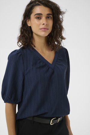 Sieviete ar cirtainiem tumšiem matiem, tērpusies tumši zilā faktūrētā V veida kakla blūzē ar pūkainām piedurknēm un melnās biksēs ar jostu.