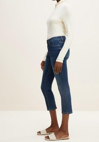 Vit långärmad topp kombinerad med högmidjade mörkblå jeans och beige tofflor, med cropped fåll och figurnära design. Slät textur.