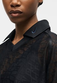 Κοντινό πλάνο ενός ατόμου που φοράει μαύρο τοπ Nike με σχέδιο δαντέλας και μαύρο γιακά με διπλωμένο μέρος με ραμμένο μπλε λογότυπο Nike.