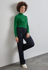 Un pull col roulé vert à rayures, un jean large bleu foncé, et des baskets blanches. Cheveux coiffés en boucles lâches. Fond uni.