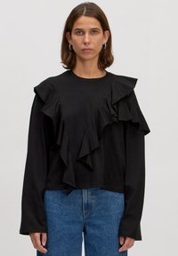 Zwarte lange mouw shirt met gelaagde ruches op één schouder, ronde halslijn en een losse fit, gecombineerd met lichtblauwe jeans.