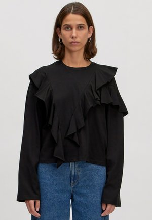 Schwarzes Langarmshirt mit gestuften Rüschen an einer Schulter, Rundhalsausschnitt und lässiger Passform, kombiniert mit hellblauen Jeans.