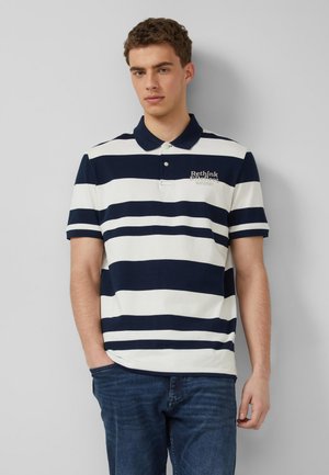 Młody mężczyzna w granatowo-białej polo z poziomymi paskami i napisem „Rethink”, stojący na tle jednolitej jasnoszarej ściany.