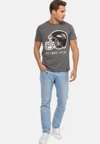 Harmaa t-paita, jossa on Baltimore Ravens -kypärän grafiikka ja teksti. Yhdistettynä vaaleansinisiin farkkuihin ja valkoisiin tennareihin, seisoo yksinkertaisella taustalla.