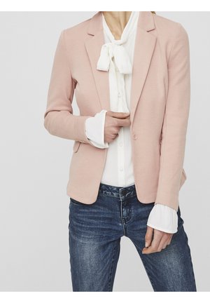 Person trägt hellrosa Blazer über weißer Bluse mit Schleife, kombiniert mit blauen Jeans, Hand passt die Vorderseite des Blazers an.