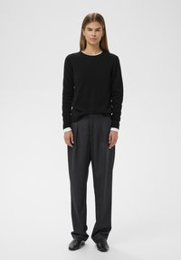 Zwarte wollen trui met een ronde hals, gecombineerd met hooggesneden grijze broek met plooien. Schoenen zijn zwart en instap. Eenvoudig, slim fit ontwerp.