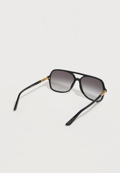 Lunettes de soleil aviateur noires avec des verres gris dégradés et des accents dorés sur les charnières, posées sur une surface gris clair.