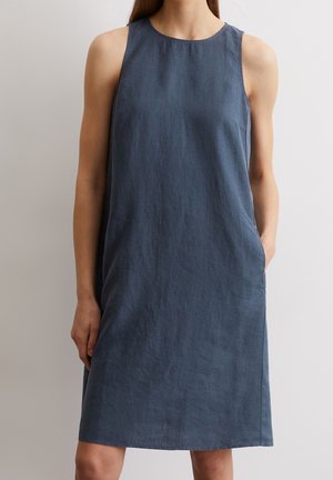 Hihaton polvipituinen tummansininen pellavamekko henkilöllä, jolla on toinen käsi taskussa, yksiväristä vaaleaa seinää vasten.