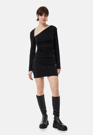 Jeune femme portant une robe mini noire ajustée à une épaule avec manches longues, subtilement scintillante, et des bottes noires épaisses montantes jusqu'aux genoux.