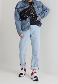 Ljusblå denimjacka över en svart grafisk T-shirt, kombinerad med avslappnade ljusa jeans och vita sneakers med röda och blå detaljer.