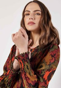 Bunt gemusterte Bluse mit gerüschten Ärmeln in Grüntönen, Orange und Rot, die an den Bündchen mit roten Rüschen akzentuiert ist.