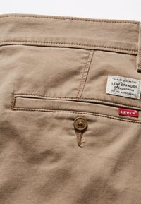 Bež pamučne hlače s stražnjim džepom, zatvaranjem na dugme i Levi's oznakom. Tekstura tkanine s detaljima ušivanja i kontrastnom niti.