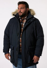Mörkblå parka-jacka med foder i päls, flera fickor och dragkedjestängning, lagd över en rutiga skjorta för extra värme och stil.