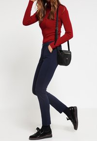 Pantalon marine ajusté et pull rouge à côtes. Un sac noir porté en bandoulière et des chaussures noires brillantes avec un détail nœud complètent le look.