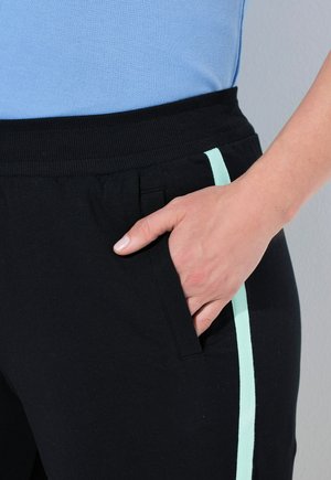 Czarne spodnie dresowe o gładkiej fakturze, z miętowo-zielonym paskiem po bokach i elastycznym pasem. Ręka w kieszeni dla skali.