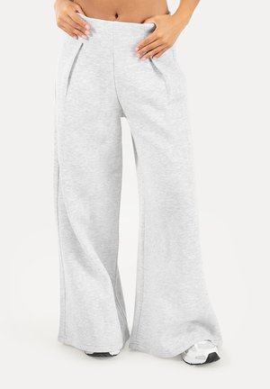Person iført lysegrå vidde joggingbukser og hvide sneakers, hænder placeret nær linningen, stående mod en ensfarvet baggrund.