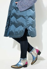 Gepolsterte türkisfarbene Daunenjacke mit einem seitlichen Schlitz, kombiniert mit metallischen Knöchelstiefeln in Blau, Silber, Pink und goldenen Akzenten, getragen über dunklen Jeans.