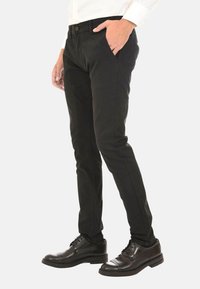 Pantaloni neri slim-fit realizzati in tessuto testurizzato con tasche laterali, indossati con scarpe eleganti nere. Chiusura a bottone visibile in vita.
