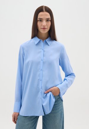 Mujer de cabello largo y oscuro que lleva una camisa de botones azul claro con rayas blancas y jeans azules, con una mano en el bolsillo.