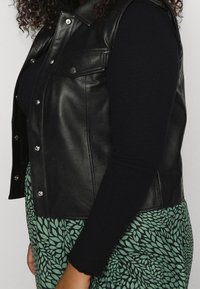 Gilet en cuir noir avec des boutons-pression en argent, associé à un haut noir à manches longues et une jupe à motif feuilles vertes. Texture lisse, design ajusté.