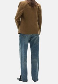 Personne portant un blazer marron et un jean droit bleu clair, debout devant un fond blanc uni.