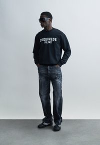 Μαύρο φούτερ με το λευκό κείμενο "DSQUARED2 MILANO", σε συνδυασμό με σκουρόχρωμα μπλε τζιν με φθορές. Το μοντέλο φοράει υπερμεγέθη μαύρα γυαλιά ηλίου και μαύρα παπούτσια.