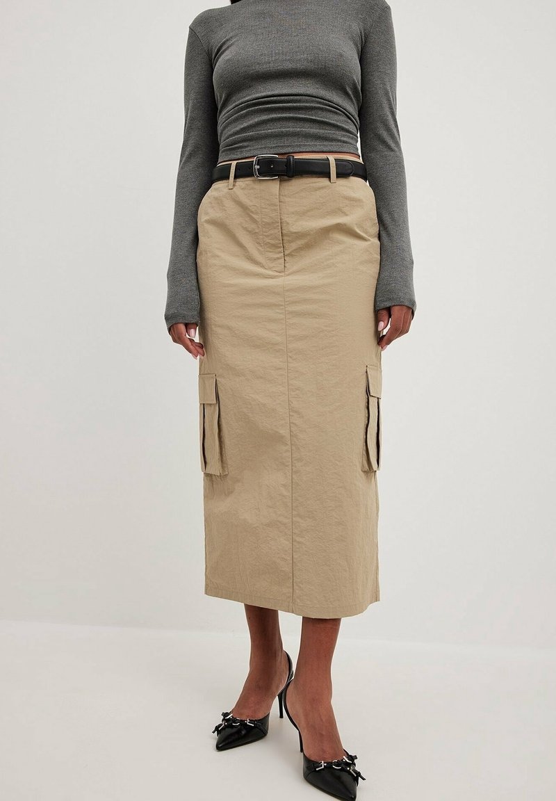 Femme portant une jupe midi beige avec poches latérales, une ceinture noire, un top ajusté gris à manches longues et des escarpins noirs pointus.