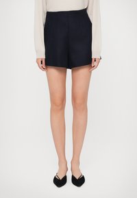 Shorts blu navy su misura con una vita alta strutturata, tasche laterali e cuciture sartoriali, abbinati a ballerine nere a punta.