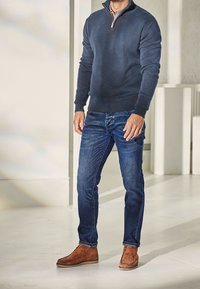 Mann in einem marineblauen Pullover mit Viertelreißverschluss, karierter Hemd, blauen Jeans und braunen Wildleder-Stiefeletten, der drinnen auf einem hellen Boden steht.
