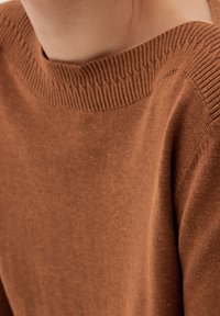 Brauner Strickpullover mit geripptem Ausschnitt und strukturiertem Stoff. Nahaufnahme des Schulter- und oberen Brustbereichs, die das Material hervorhebt.