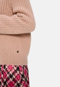 Helles rosa gestricktes Sweatshirt mit gerippter Textur, das ein kleines Logo-Detail aufweist. Kombiniert mit einem bunt gemusterten Rock in Pink und Schwarz.