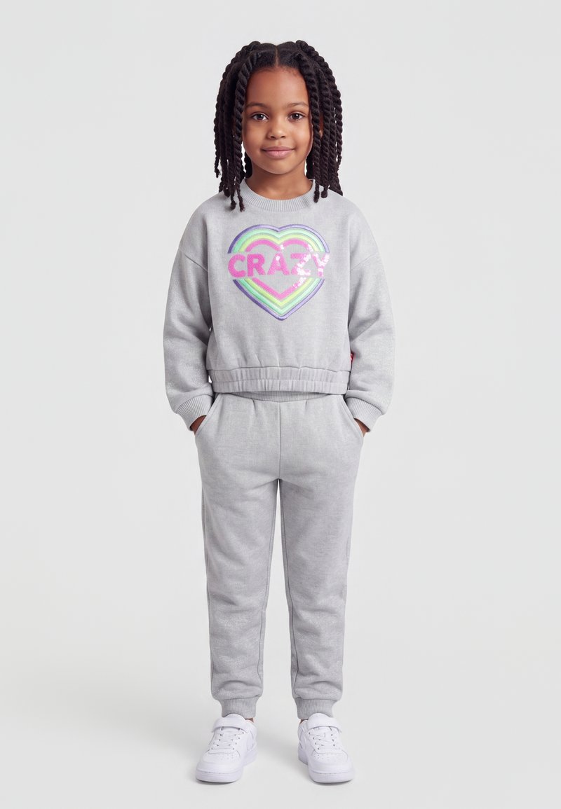 Jeune fille avec des tresses portant un sweat-shirt gris et un pantalon avec un cœur coloré et le mot "CRAZY,", debout les mains dans les poches.