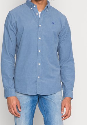 Mężczyzna ubrany w jasnoniebieską koszulę z długim rękawem zapinaną na guziki, na białej koszulce pod spodem, w połączeniu z jasnoniebieskimi dżinsami.