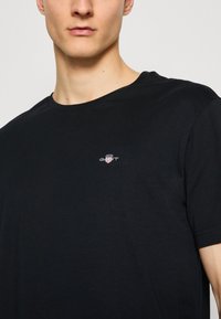 T-shirt en coton noir avec un col rond, des manches courtes, et un petit logo comportant des rayures rouges et blanches sur la poitrine.