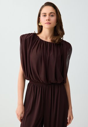 Vrouw met schouderlang bruin haar, die gouden oorbellen draagt en een donkerbruin mouwloos geplooid top- en broekset, staand tegen een effen achtergrond.