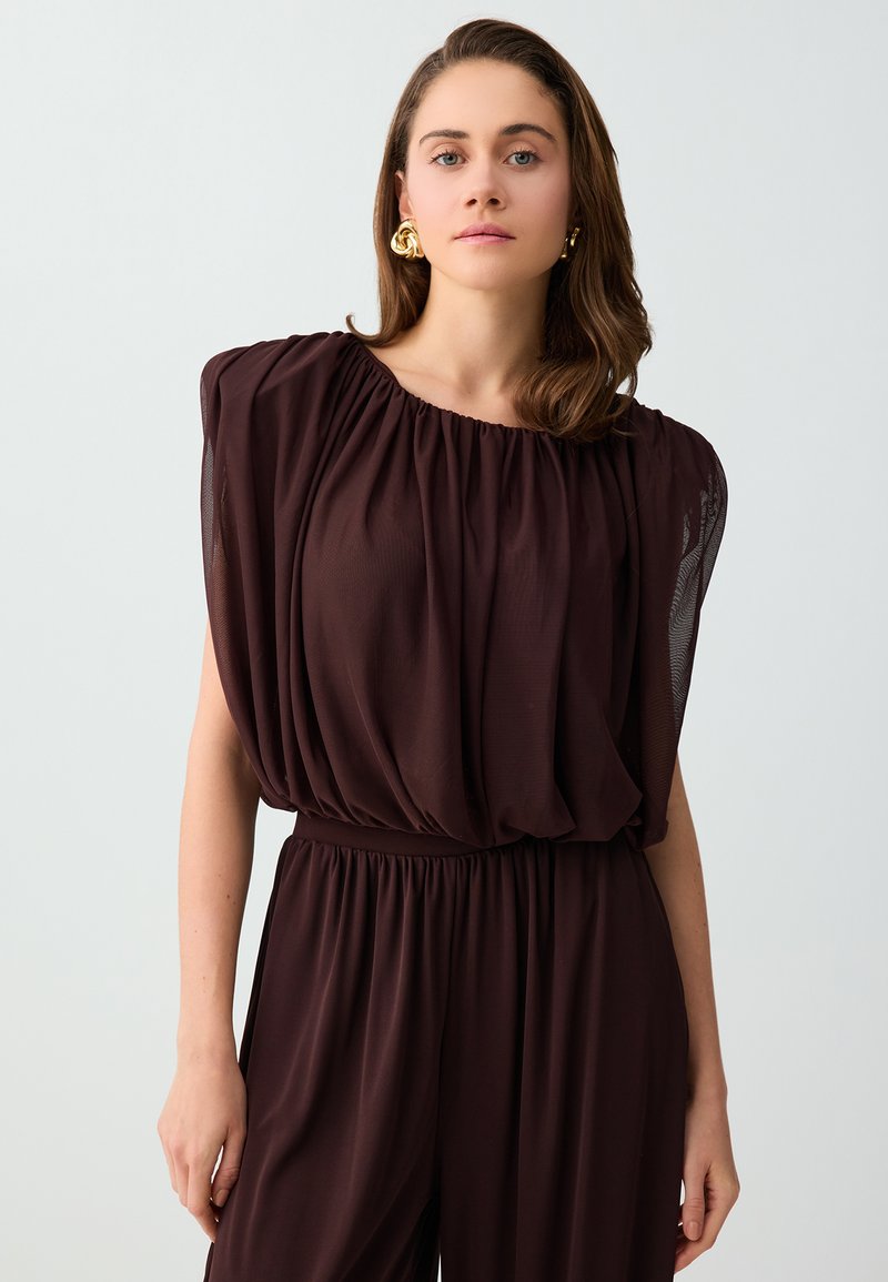 Femme aux cheveux bruns mi-longs, portant des boucles d'oreilles en or et un ensemble top sans manches plissé et pantalon marron foncé, debout devant un fond uni.