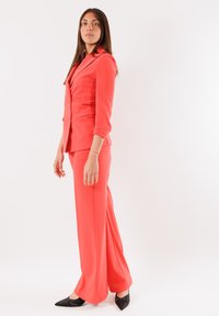 Completo in corallo su misura con maniche lunghe, giacca aderente e pantaloni a gamba larga, abbinato a scarpe nere con punta affilata. Tessuto di texture liscia.