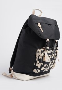 Mochila de tela negra con acentos crema, correas ajustables y un bolsillo frontal con patrón. Cuenta con una solapa superior y cierre de cremallera.