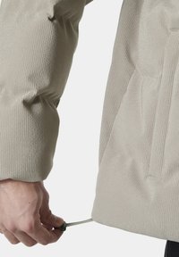 Lichtbeige textuurjasje met een geribbeld ontwerp. Hand die de koordjes aan de onderzoom aanpast. Heeft zijzakken en een ritssluiting.